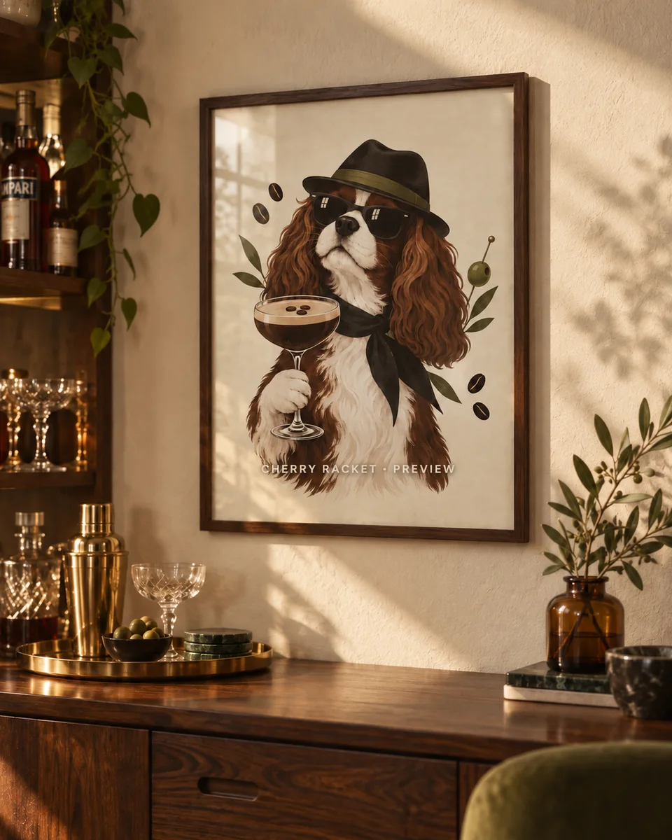 Room mockup: a framed illustration of a spaniel in sunglasses holding an espresso martini, on a warm plaster wall above a dark wood sideboard.